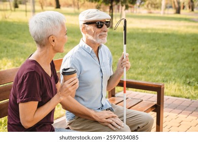 Side view of elderly couple sitting on a bench in park sunny day. Old blind man with walking stick and woman drinking coffee talking outdoors. Compassion caregiving life in retirement concept