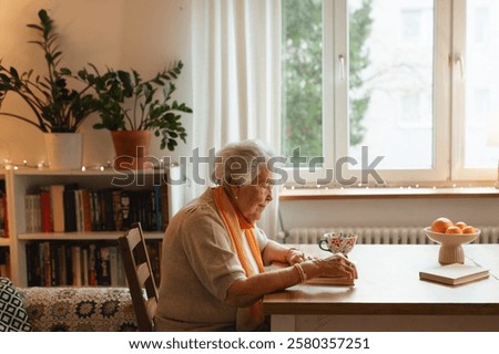 Side view of elderly lady reading a book, sitting at the kitchen table and sipping tea.
