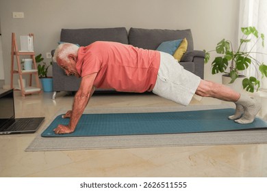 Side view of elderly man doing plank exercise on a yoga mat following online fitness class on laptop at home - Powered by Shutterstock - Get 15% off with code: PIKWIZARD15