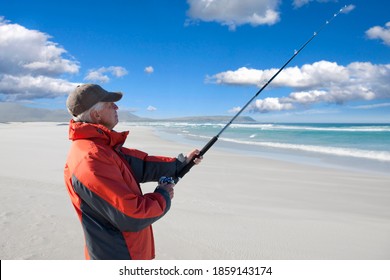 Side View Of An Elderly Man Fishing At The Shore Of A Sunny Beach.