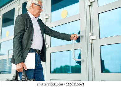 Side View Of Elderly Man With Laptop And Briefcase Opening Door Of Office Building While Walking To Work