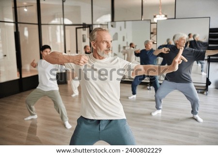 Side view of elderly people in gym, focus on bearded senior man posing as archer shooting during qigong lesson in gym