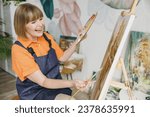 Side view elderly smiling artist woman 50 years old wear casual clothes sitting near easel with painting artwork paint hold palette spend free spare time in living room indoor. Leisure hobby concept