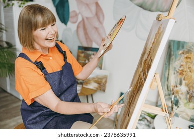 Side view elderly smiling artist woman 50 years old wear casual clothes sitting near easel with painting artwork paint hold palette spend free spare time in living room indoor. Leisure hobby concept - Powered by Shutterstock