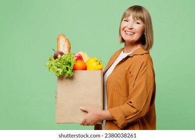 Side view elderly smiling happy positive woman wear brown shirt casual clothes hold shopping paper bag with food products looking camera isolated on plain green background. Delivery service from shop