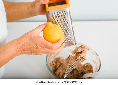 Side view of an elderly woman's hand rubbing lemon peel into cup of cream pie filling, inside room. Retired housewife cooking hobbies in kitchen. Selective focus on fruit.