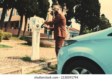 Side view of an elegant African-American mature guy with a black beard and in glasses plugging his car into an outdoor charger; a dapper African man is connecting his vehicle to a street power supply