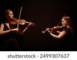 Side view of elegant string duet of female violinists playing classical music in contrast top light on stage with black curtains, copy space