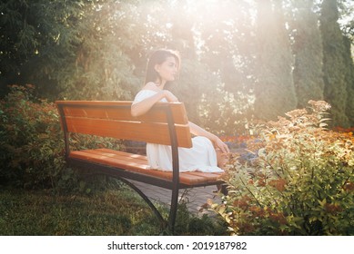 Side view of elegant young brunette woman in white dress resting on bench in park in rays of sun, enjoying weekend outdoors. Beautiful girl with dark skin sitting on bench and looking to the side. - Shutterstock ID 2019187982