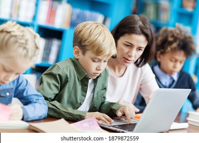 Side View Of Elementary Student Doing Task On Laptop Computer Helped By Female Teacher, His Defocused Classmates Writing In Notebooks Sitting At Desk In School Classroom Nearby