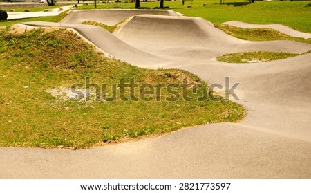 Side view of an empty asphalt pump track featuring undulating humps, berms, and curves integrated into a green public park. Perfect for commercial marketing, sports branding, outdoor recreation campaigns, youth activity promotion, cycling, skateboarding culture, and modern eco-friendly urban space concepts.