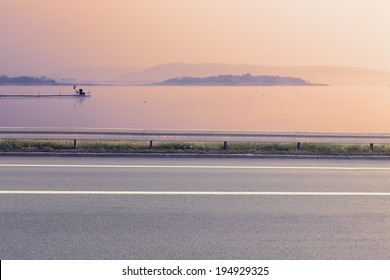 Side View Of Empty Asphalt Road And Lake At Sunrise