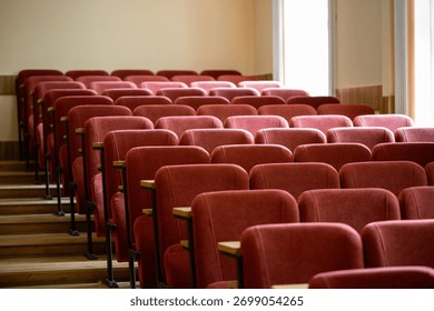 A side view of an empty university lecture hall. Rows of maroon seats with small wooden desks are arranged on stepped risers, with bright windows in the background.