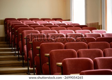A side view of an empty university lecture hall. Rows of maroon seats with small wooden desks are arranged on stepped risers, with bright windows in the background.