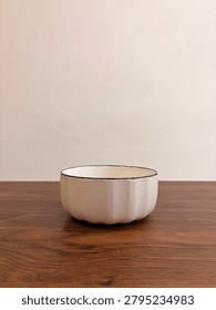 Side view of an empty white ceramic bowl with a black rim placed on a wooden table against a neutral background. 