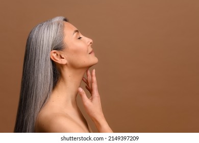 Side View At Enchanting Topless Middle Aged Asian Woman Isolated On Brown Background, Charming Korean Lady With Grey Hair Stands In Profile And Touching Chin Gently. Skin Care And Self Love Concept