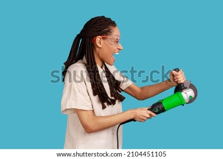 Side view of enthusiastic woman with dreadlocks working with grinder saw, yelling with excitement, wearing white shirt and protective glasses. Indoor studio shot isolated on blue background.