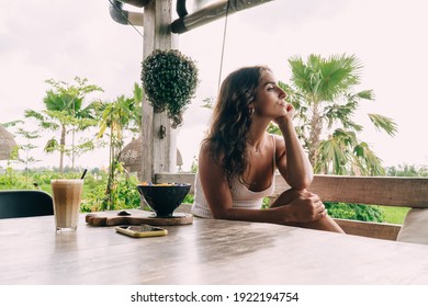 Side View Of Ethnic Female Leaning On Hand While Sitting At Table With Breakfast Bowl And Coffee In Outdoor Cafe In Tropical Resort