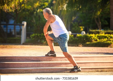 Side View European Bearded Old Man In White Vest Blue Shorts Does Morning Exercises Squats On Right Knee On Stone Steps
