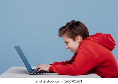 Side View Excited Male Teenager Red Sweatshirt Playing Game On Laptop At Table In Studio On Blue Background