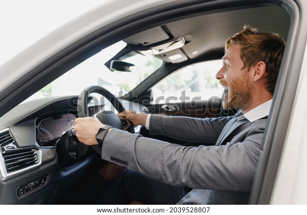 Side view excited man customer buyer client in\
classic grey suit driving car hold wheel choose auto want buy new\
automobile in showroom vehicle salon dealership store motor show\
indoor Sale concept