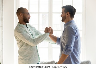 Side View Excited Mixed Race Male Friends In Eyewear Shaking Hands. Happy Best Buddies Welcoming Each Other At Sudden Meeting. Smiling Coworkers Saying Hi, Communicating Together Indoors.