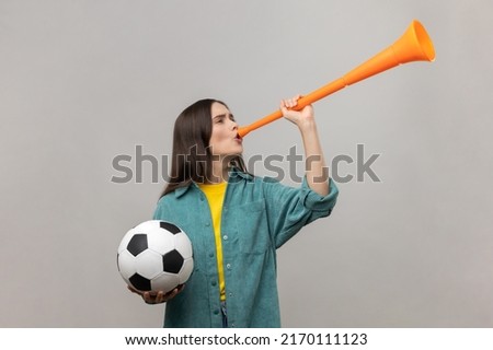 Side view of excited woman blowing in horn holding black and white soccer ball, celebrating victory of favourite football team, wearing casual jacket. Indoor studio shot isolated on gray background.