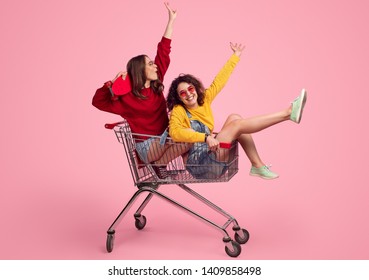 Side view of excited young friends smiling and raising hands while riding shopping trolley against pink background - Shutterstock ID 1409858498