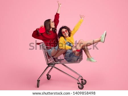 Side view of excited young friends smiling and raising hands while riding shopping trolley against pink background