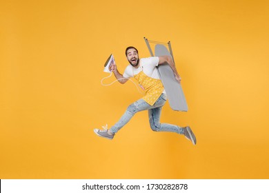 Side view of excited young man househusband in apron rubber gloves hold iron board for ironing while doing housework isolated on yellow background studio. Housekeeping concept. Jumping looking camera