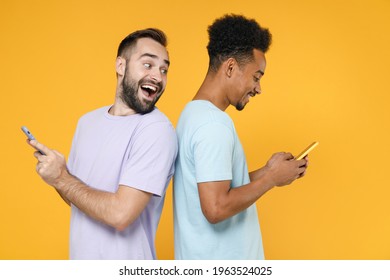 Side View Of Excited Young Two Friends European African American Men 20s Wearing Violet Blue Casual T-shirts Using Mobile Cell Phone Typing Sms Message Isolated On Yellow Background Studio Portrait