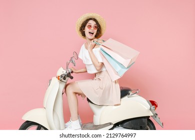 Side view of excited young woman 20s wearing white summer clothes hat glasses hold package bag with purchases after shopping driving moped isolated on pastel pink colour background studio portrait