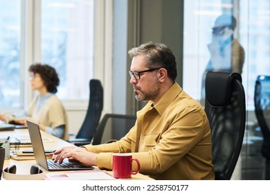 Side view of experienced manager typing on laptop keypad and looking at screen by workplace while networking in openspace office