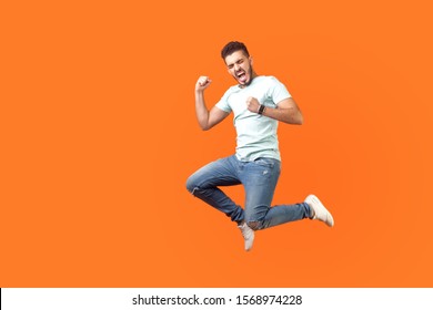 Side view of extremely happy man in white outfit jumping in air with raised arm gesturing yes i did it, I'm winner, screaming loud for joy feeling energetic and lively. isolated on orange background