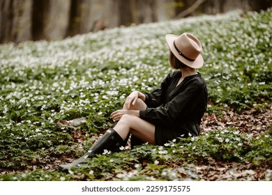 Side view of faceless woman resting on grass in forest. Girl in hat, dressed in black dress and rubber boots, enjoying nature in the blooming forest with white flowers - Shutterstock ID 2259175575