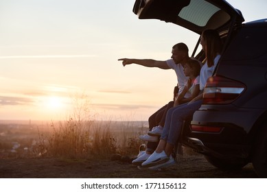 Side View Of The Family Sitting In The Car Trunk Outside The City On The Hill Watching The Sunset, Father Is Pointing On The Horizon, Copy Space