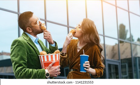 Side View Of Fashionable Couple In Velvet Clothing Eating Fried Chicken Legs On Street