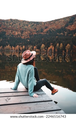 Side view of fashioned young woman sitting on wooden dock looking at view with beautiful autumn colors. Female hipster with brown hat relaxes on the edge of jetty. Wonderful nature getaway
