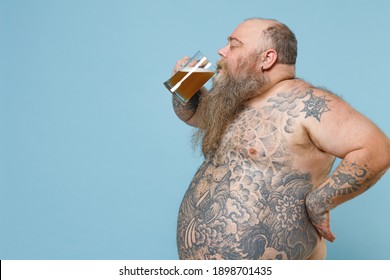 Side view of fat pudge obese chubby overweight man has tattooed naked bare big belly hold glass of beer isolated on blue color background studio portrait. Weight loss obesity unhealthy diet concept