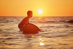 Side View Of Fearless Kid Floating Surfboard At Ocean With Waves On Sunny Evening. Surfer With His Surfboard At The Beach