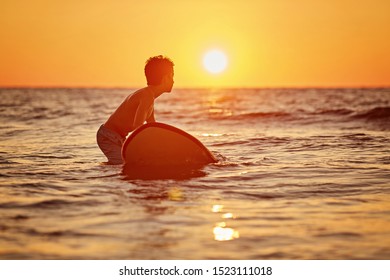 Side View Of Fearless Kid Floating Surfboard At Ocean With Waves On Sunny Evening. Surfer With His Surfboard At The Beach