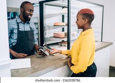 Side View Of Female African American Customer Using Mobile Phone And Paying With NFC Technology In Confectionery Shop While Salesman Standing At Counter With Payment Terminal