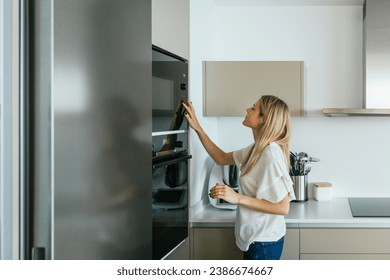 Vista lateral de la mujer con un traje informal que da la vuelta al moderno horno microondas negro mientras está de pie en la cocina con utensilios modernos de cocina