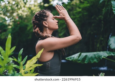 Side view of female with closed eyes sitting among green plants with raised hands in Namaskar Mudra and touching both thumbs to forehead and meditating in green park - Powered by Shutterstock