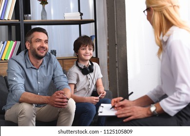 side view of female counselor writing in clipboard while father and son with headphones sitting on therapy session in office 