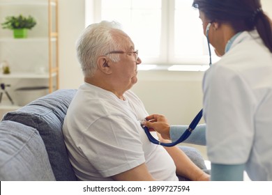 Side View Of A Female Doctor Listening To The Heart And Breathing Of An Older Man Through A Stethoscope On His Chest. Man Sitting On Sofa At Home. Concept Of Medical Care For The Elderly At Home.