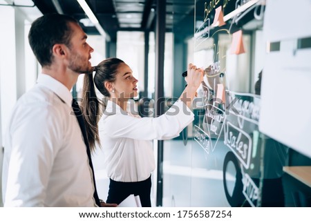 Side view of female drawing scheme of business strategy on glass board while male coworker observing process at modern workplace