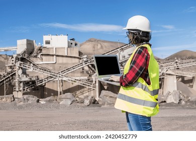 Side view of female engineer in hardhat and waistcoat working on laptop with black screen while visiting construction site of unfinished industrial facility