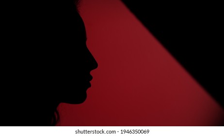 Side view of female face placing her finger on lips on dark red background. Closeup silhouette of serious woman showing hush gesture. Faded hand making keeping secret sign.