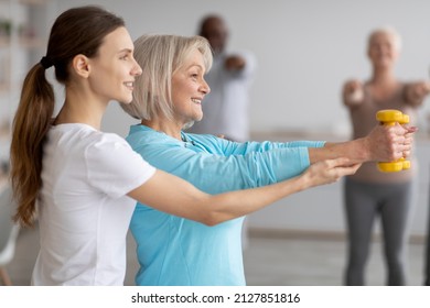 Side View Of Female Fitness Coach Helping Active Senior Woman While Exercising With Dumbbells, Sporty Multiracial Group Of Elderly People Having Fitness Class At Nursing Home, Training With Instructor